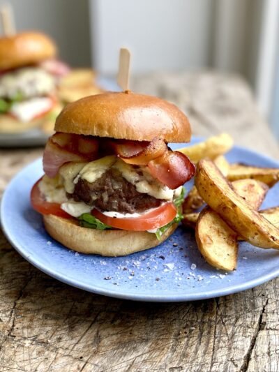 Two burgers sitting on blue plates and serve with melted cheese and bacon with sliced tomatoes and a side of potato wedges
